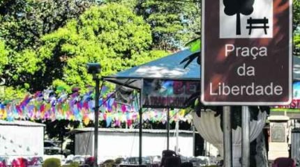 Bandeirolas enfeitam a praça para quermesse. Frequentadores apreciam vegetação conservada do espaço