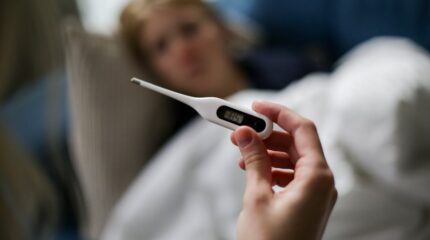 Mão segurando termômetro digital com pessoa doente desfocada ao fundo.